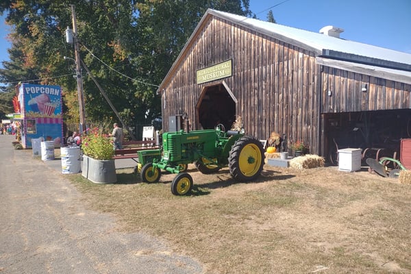 Weathered wooden Agricultural Museum barn at the Farmington Fairgrounds with a green John Deere tractor out front and a popcorn stand to the left